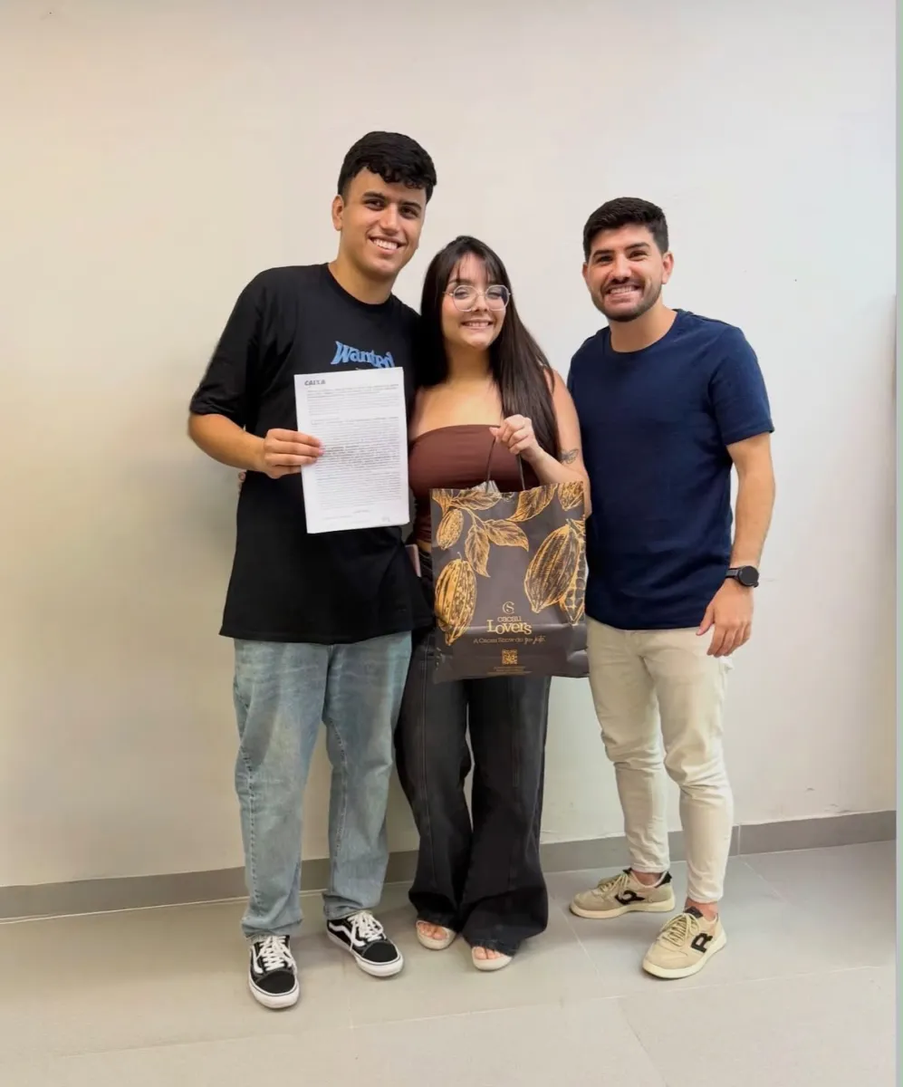 Casal com Matheos em ambiente interno de parede branca, o rapaz segura o contrato assinado; a moça, de óculos, carrega uma sacola de presente de chocolateria.