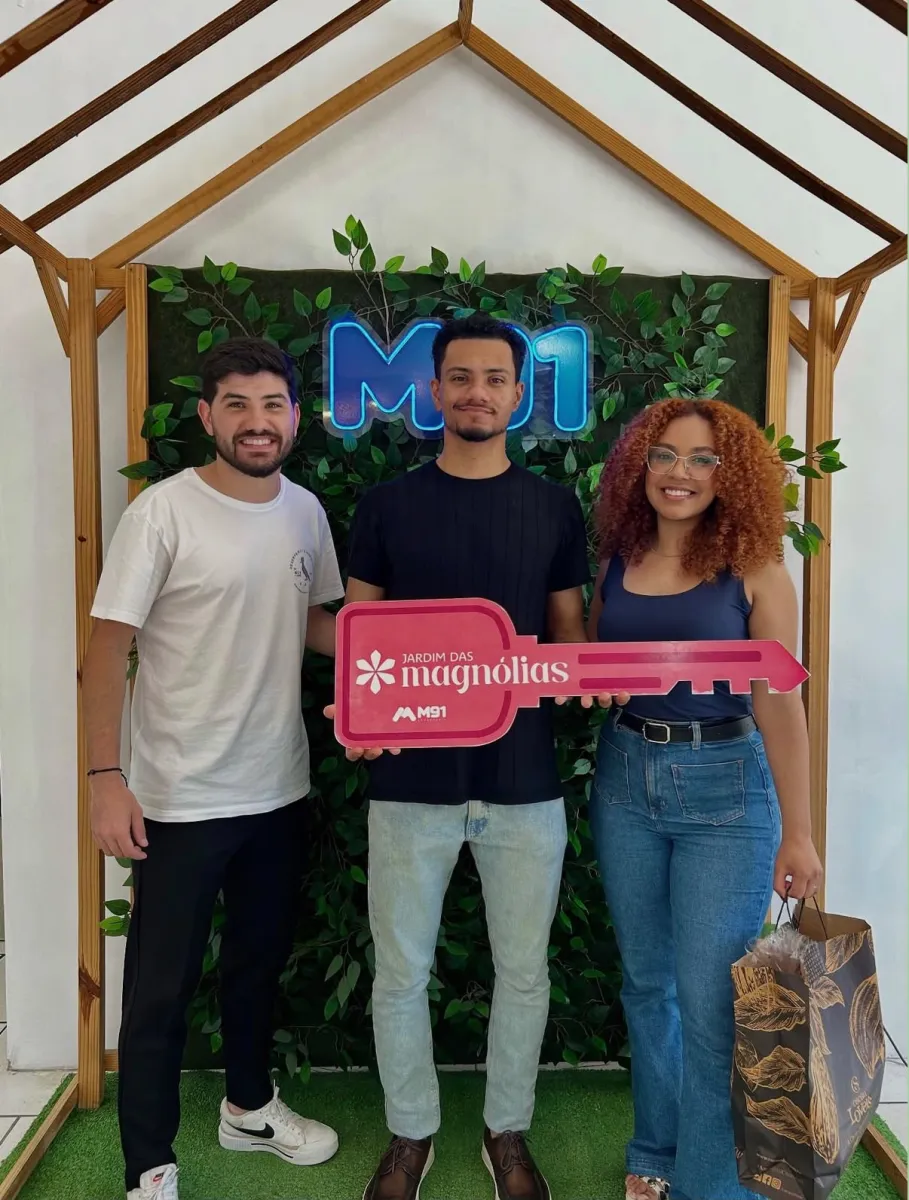 Trio diante de painel decorado com folhagem, o homem do meio segura uma placa comemorativa cor-de-rosa em formato de chave; a mulher à direita, de cabelo cacheado e óculos, sorri ao lado dele.