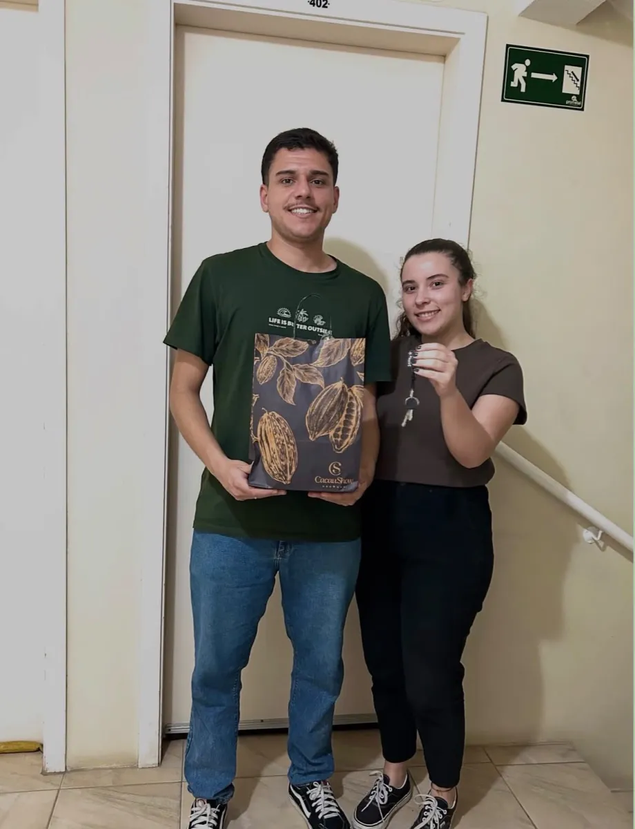 Casal em corredor de prédio, parado diante de uma porta numerada, o homem segura uma sacola de presente de chocolateria enquanto a mulher mostra um molho de chaves reais na mão.