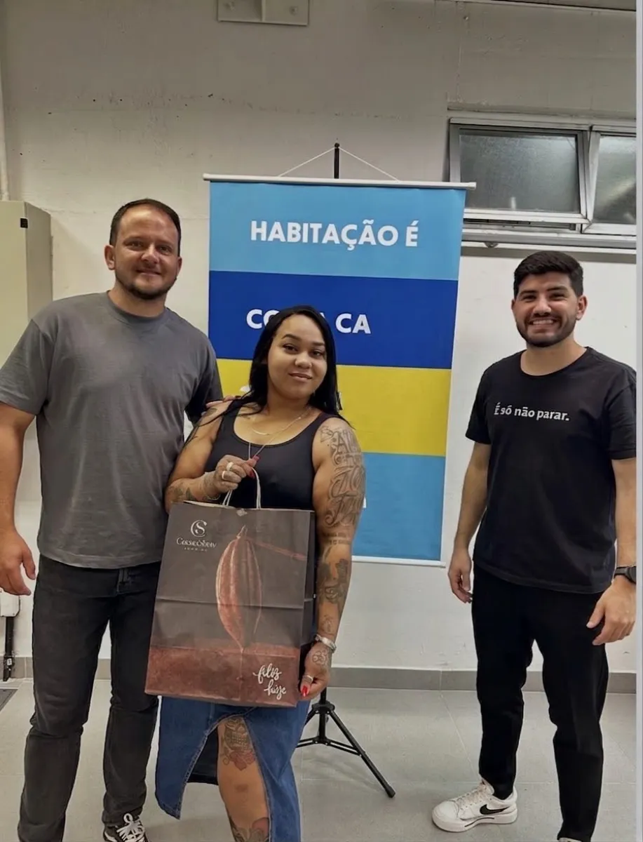 Trio diante de um banner sobre habitação, a mulher no centro, com tatuagens no braço, carrega uma sacola de presente de chocolateria entre os dois homens ao seu lado.